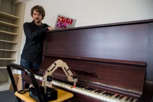 Marius Schmeding lehnt am Klavier und sieht dem Roboterarm beim Spielen zu. Foto: Holger Vogel